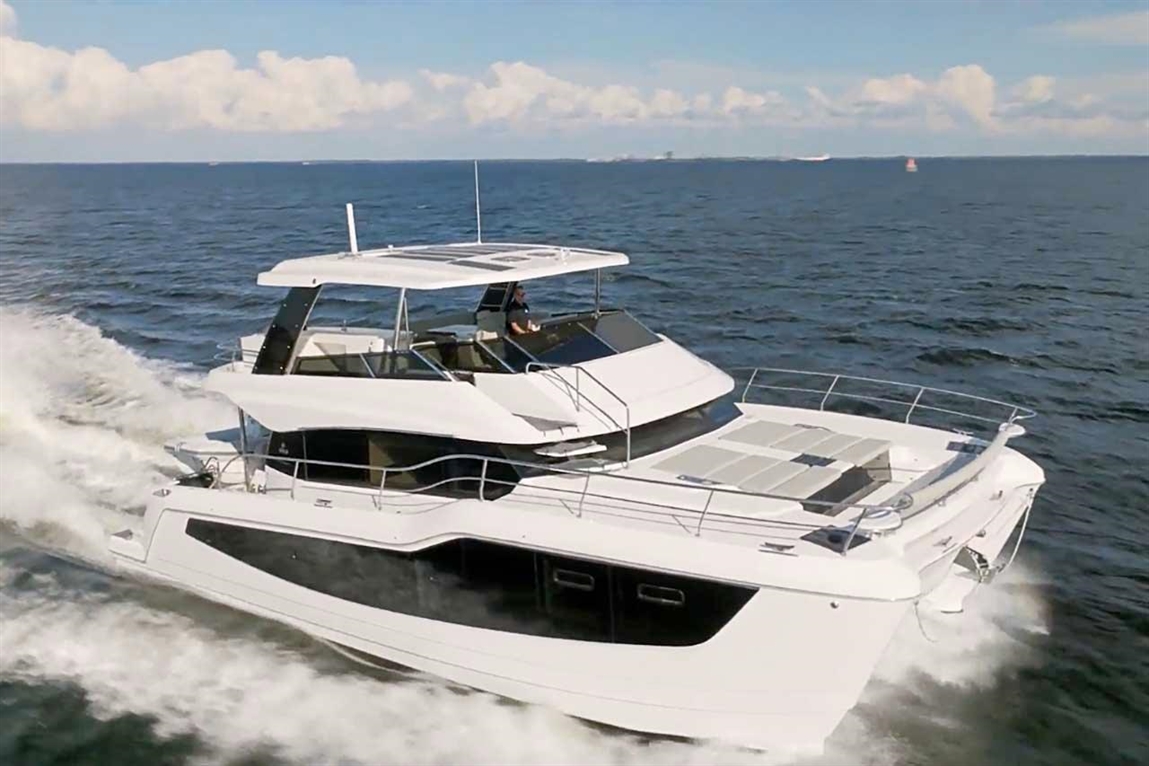 Luxury motor yacht cruising on blue waters with a captain at the helm, surrounded by clouds and distant boats.